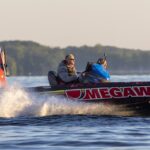 Two people ride a high-speed bass boat with "MEGAWARE" on the side, creating a spray of water against a backdrop of calm water and distant trees