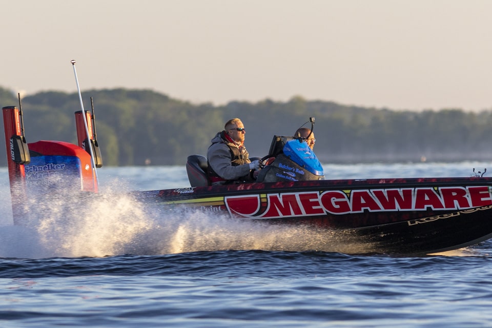 Two people ride a high-speed bass boat with "MEGAWARE" on the side, creating a spray of water against a backdrop of calm water and distant trees