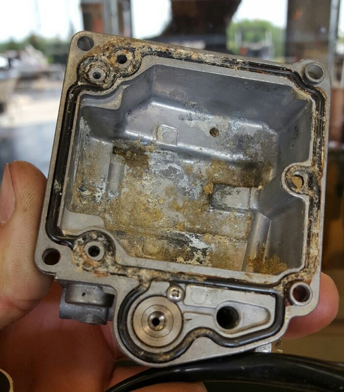 A person's hand holds a corroded and tarnished metal carburetor part with visible sealant residue inside